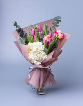 Handmade bouquet for Mother's Day in a festive paper package stands in a vase on a blue background. Beautifully packaged flower arrangement with pink tulips, white hydrangea and eucalyptus twigs.