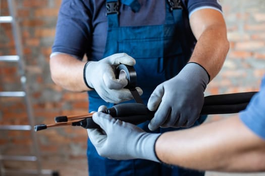 Two adult male workers in overalls are preparing to install underfloor heating pipes. Close up of repairmen hands in work gloves fastening pipes and wires with insulating tape.