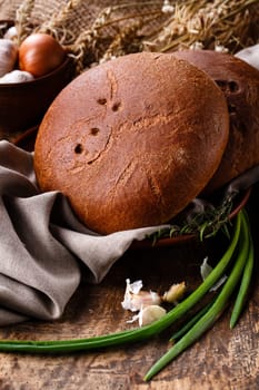 Two fresh loaves of bread on clay plate on table with rustic natural elements. Close-up of baked round black bread lies on table on side of which lie burlap, garlic and green onions.