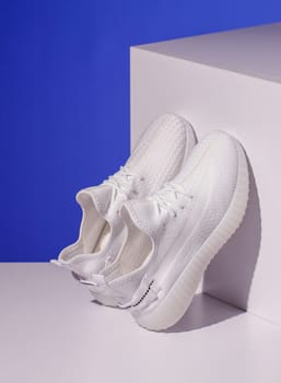 Pair of white sneakers stand on white geometric podium on blue background. Women's stylish sneakers stand leaning on cube. Spring and summer fashion concept.