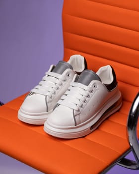 Close up of white sneakers standing on red chair on purple background in studio. Sneakers are very popular among teenagers or women because of their simple and fashionable appearance.
