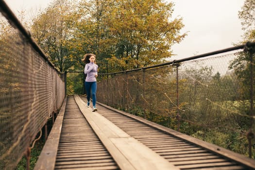 Young woman spends time with benefit. Female runner begins to jog over a wooden suspension bridge in the middle of a mountain forest. Fitness woman working out in nature on a cool autumn day.
