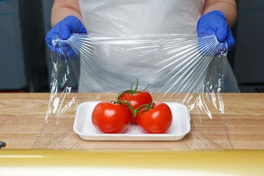 Grocery retailers. Packing of food products. Unknown worker in blue disposable gloves wraps in food transparent film tomatoes lying on white plastic tray. Food delivery concept.