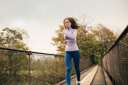 Living healthy lifestyle. Young sporty woman running across a suspension bridge during cardio exercises. Female fitness athlete practicing jogging outdoors. Jogging, active lifestyle concept.