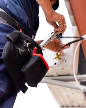 Male technician connects outdoor air conditioning unit to wall of old apartment building. Close-up of worker pulling tool out of his waist bag. Selective fiocus.