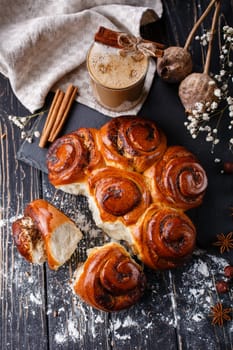 Freshly baked cinnamon buns with spices and stuffing with coffee drink on wooden table. Close up of buns on black methal board on table near Christmas spices. Christmas baking concept. Top view.