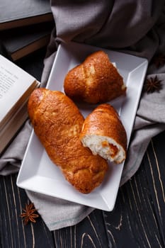 Homemade pastries. Freshly baked fragrant buns with jam inside lie near open book on wooden table. Top view of one whole bun and one sliced lying on white plate next to burlap, books, and star anise.