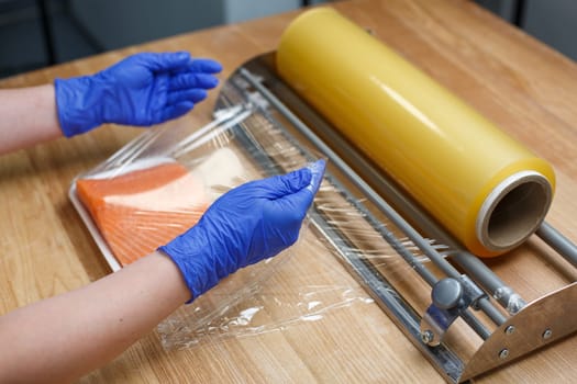 Worker in blue disposable gloves wraps piece of salmon fillet lying on white plastic tray in transparent film. Concept of storage, keeping salted and raw fish and delivery to customer. Close up.