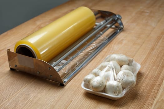 Close up of champignons on plastic tray packed in transparent cling film. Fresh packaged mushrooms on table near large roll of food packaging film. Concept of storing fresh food in supermarkets.