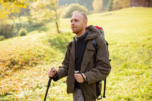 Sporty tourist man with trekking poles and a backpack travels through the mountain hills. Smiling man enjoys the beauty of nature. Concept of hiking, healthy and active lifestyle in young people.