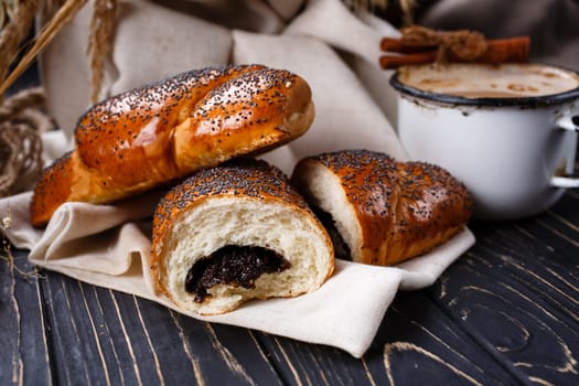 Freshly baked fragrant homemade buns with jam on rustic wooden table. Close up of cut buns sprinkled with poppy seeds and lying on sackcloth near enameled cup of coffee.