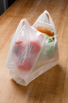 Close up of plastic bag with various vegetables inside which are wrapped in cling film. Grocery package from supermarket or grocery store stands on wooden table. Concept of using lot of plastic.
