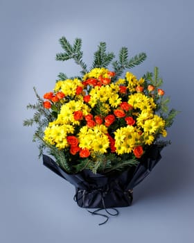 Original bouquet of small pink roses, yellow chrysanthemums and spruce twigs. Bouquet wrapped in black floral paper with ribbon standing on a blue background. Funeral bouquet