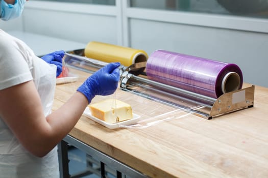 Food packaging. Woman in gloves, apron and mask wraps piece of cheese lying on plastic tray in transparent food film. Concept of food packaging in supermarkets or suppliers of healthy food.