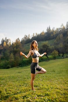 Yoga outdoor. Cute slender young woman with closed eyes meditates, does breathing exercises and keeps balance on one leg standing barefoot on the grass outdoors. Concept of physical and mental health.