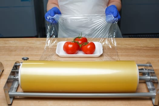 Grocery retailers. Packing of food products. Unknown worker in blue disposable gloves wraps in food transparent film tomatoes lying on white plastic tray. Food delivery concept.