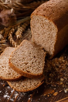 Homemade baking. Close up of sliced rye bread on dark wooden background. Freshly baked fragrant brown bread lies on wooden table on background of ears of rye or wheat. Selective focus.