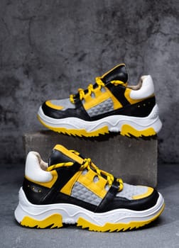 Pair of stylish bright sneakers in yellow, black and white on background of gray concrete. For better visualization one sneaker stands on stone pedestal and other on floor. Selective focus.