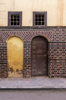 Stone blocked arced door, decorated wooden arched door, and two small windows with iron rusted bars on black and red bricks wall on cobblestone road