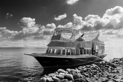 Black and white shabby aged boat floating in sea water near rocky coast against cloudy sky