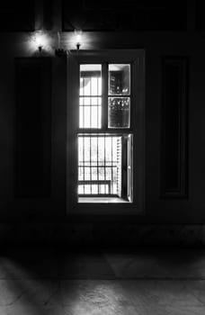 Black and white high contrast shot of narrow window, revealing strong light into dark room with illuminated lanterns and white tiled marble floor
