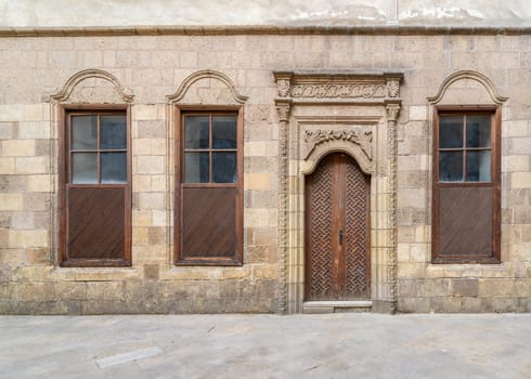 Facade of old abandoned stone decorated bricks wall with arched wooden door and three wooden shutter windows