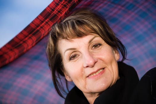 Head portrait of a mature woman in undersight in front of blue sky with a plaid headscarf waving in the wind behind her,smiling friendly at the camera