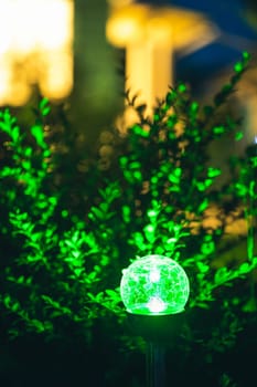Night View Of Flowerbed Illuminated By Energy-Saving Solar Powered Colorful Multi-colored Lantern On Yard. Beautiful Small Garden With Green Light, Lamp In Flower Bed. Garden Design.