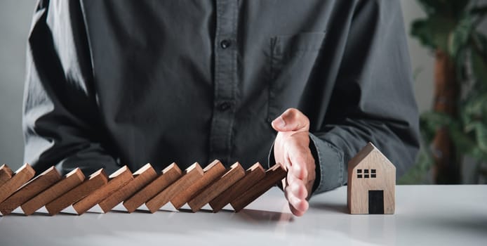 Business risk plan real estate protection. Man hand blocks wood block from many row falling wooden block like domino to house model, Insurance ideas to prevent loss concept