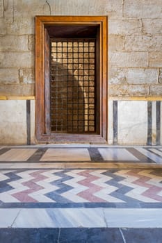 Recessed wooden window with decorated iron grid over stone bricks wall and decorative colorful floor at public historic mosque of Sultan Barquq, El Moez Street, Old Cairo, Egypt