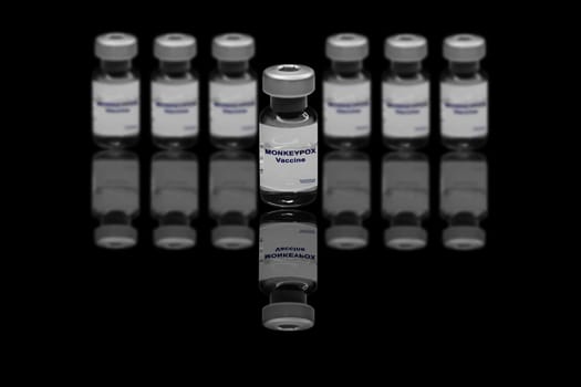 Monkeypox vaccine close-up on a black mirror background. In the background are blurred vaccine vials.