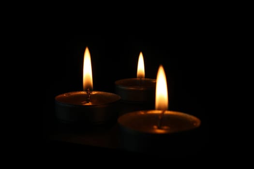 Three burning candles close-up on a black background with copy space. Blurred foreground and background.