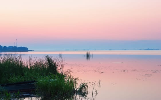 Pastel soft pink dawn on a calm lake overgrown with reeds. Blue horizon and clear sky are reflected in the smooth water surface before sunrise. Picturesque landscape with space for text. Rostov Veliky, Lake Nero.