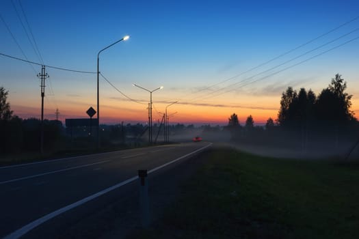 Asphalt road with markings and traces of the headlights of a moving car in the foggy countryside at sunset. Street lights and power lines along the highway. Road A-119 in Vologda Oblast, Russia.