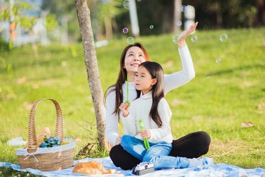 Happy Asian mother and little girl daughter child having fun and enjoying outdoor together sitting on the grass blowing soap bubbles during a picnic in the garden park on a sunny day family time