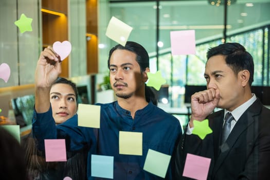 Businessman writing sticking adhesive notes on glass wall in office and team help to creative idea, Teamwork working write work stickers reminder on paper at widows, business and collaboration concept