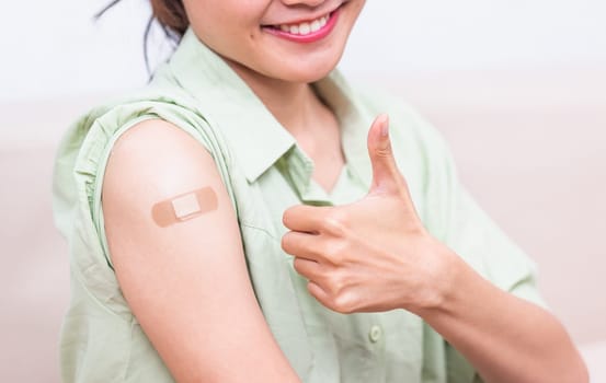 Asian young woman showing gesturing thumb up or like and her arm with plaster band aid after coronavirus Covid-19 vaccine injection on shoulder, coronavirus COVID-19 vaccination concept