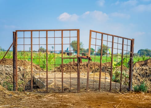 Weathered old gate tied with rope and installed near heaps of soil against cloudy sky on sunny day on farm