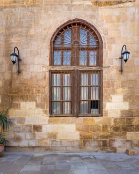 Facade of old abandoned stone bricks wall with arched wooden window covered by wrought iron bars and two lanterns, Cairo, Egypt