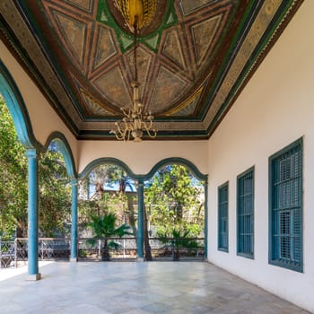 Covered terrace with decorated painted ceiling, green stone pillars, and marble floor fenced with metal forged fence and surrounded with green trees in sunny summer day