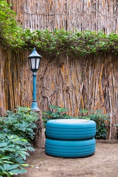 Small streetlamp and painted blue tires located near thatched fence and green plants in yard in countryside
