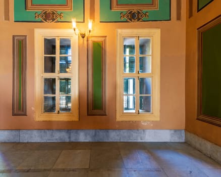 Corner orange wall with two narrow wooden window with closed green shutters, beautiful elegant rectangular green frames, and white tiled marble floor