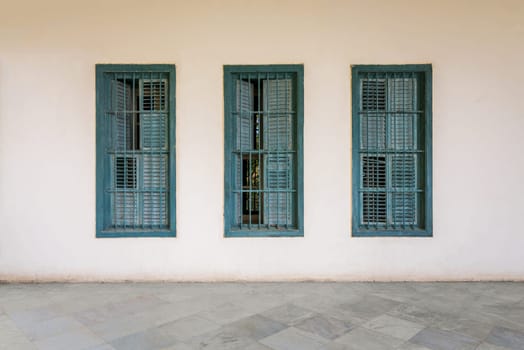 White wall with three grunge windows with wooden green shutters and wrought iron bars and white marble floor