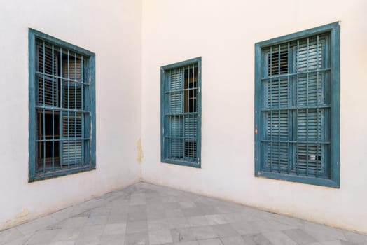 Corner white wall with three grunge windows with wooden green shutters and wrought iron bars and tiled white marble floor