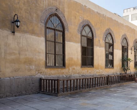 Row of three adjacent wooden grunge arched window with decorated wrought iron grid over yellow stone bricks wall and wooden balustrade, Medieval Cairo, Egypt