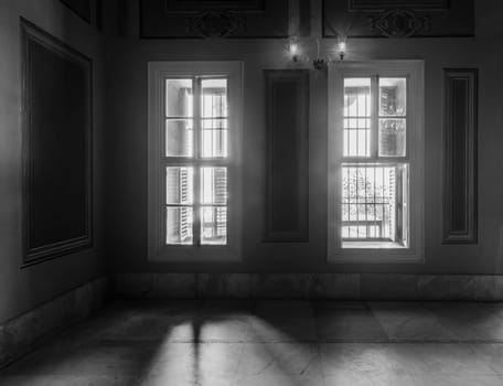 Black and white high contrast shot of two narrow windows, revealing strong light into dark room with white tiled marble floor