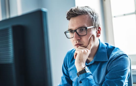Handsome Young Businessman Wearing Eyeglasses Sitting at his Table Inside the Office, Looking at the Report on his Computer Screen