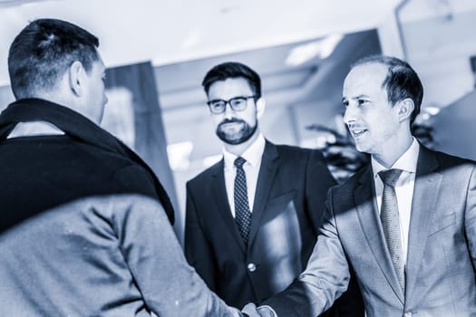 Group of confident business people greeting with a handshake at business meeting in modern office. Closing the deal agreement by shaking hands. Greyscale blue toned image.
