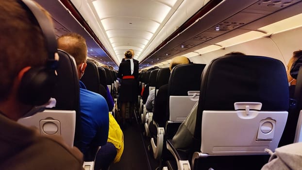 Interior of airplane with passengers on seats and stewardess in uniform walking the aisle.