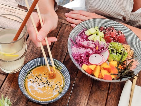 Woman eating tasty colorful healthy natural organic vegetarian Hawaiian poke bowl using asian chopsticks on rustic wooden table. Healthy natural organic eating concept.